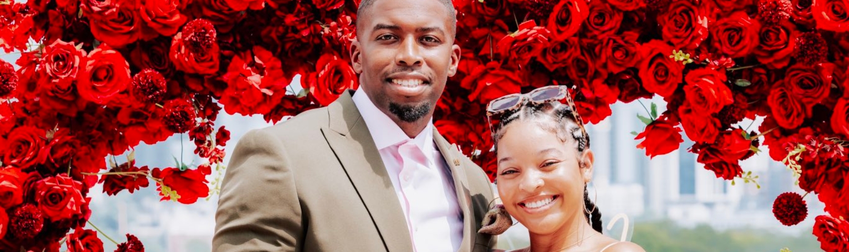 A Black man in a olive green suit stands next to a Black woman. They are in front of an arch made of rose petals.
