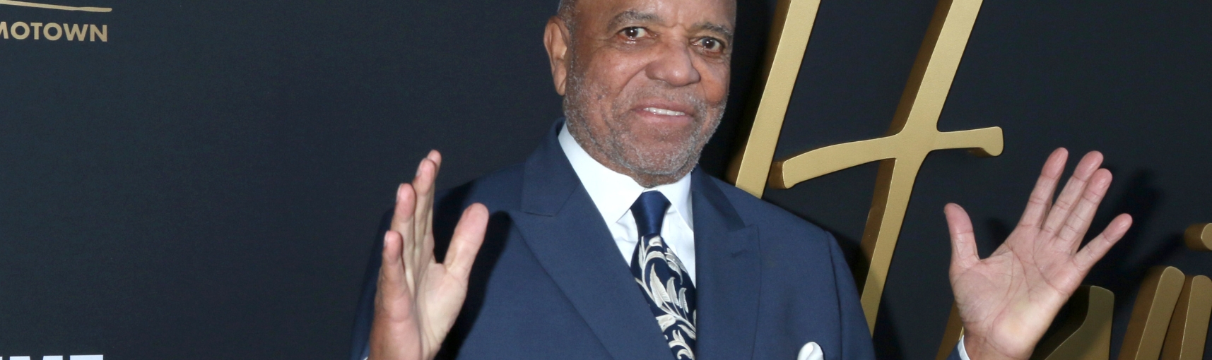 A Black man in a tuxedo smiling as he holds his hands up