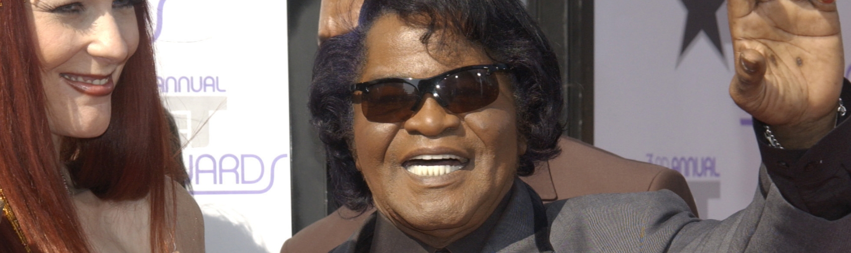A woman with red hair stands next to an older Black man with black hair and sunglasses